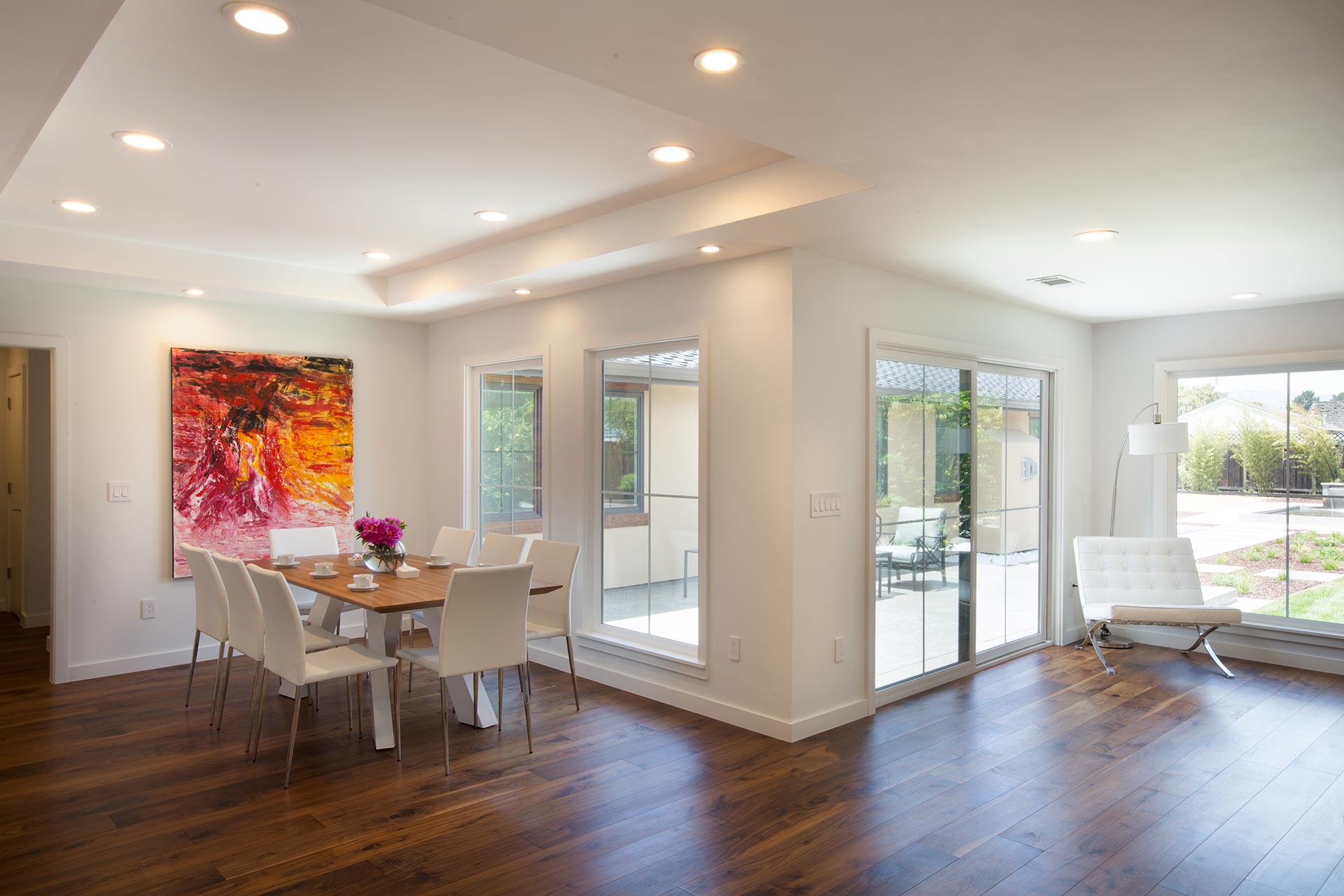 Modern dining room with wooden floor, a table with six chairs, a vibrant painting on the wall, and large windows leading to an outdoor patio with greenery